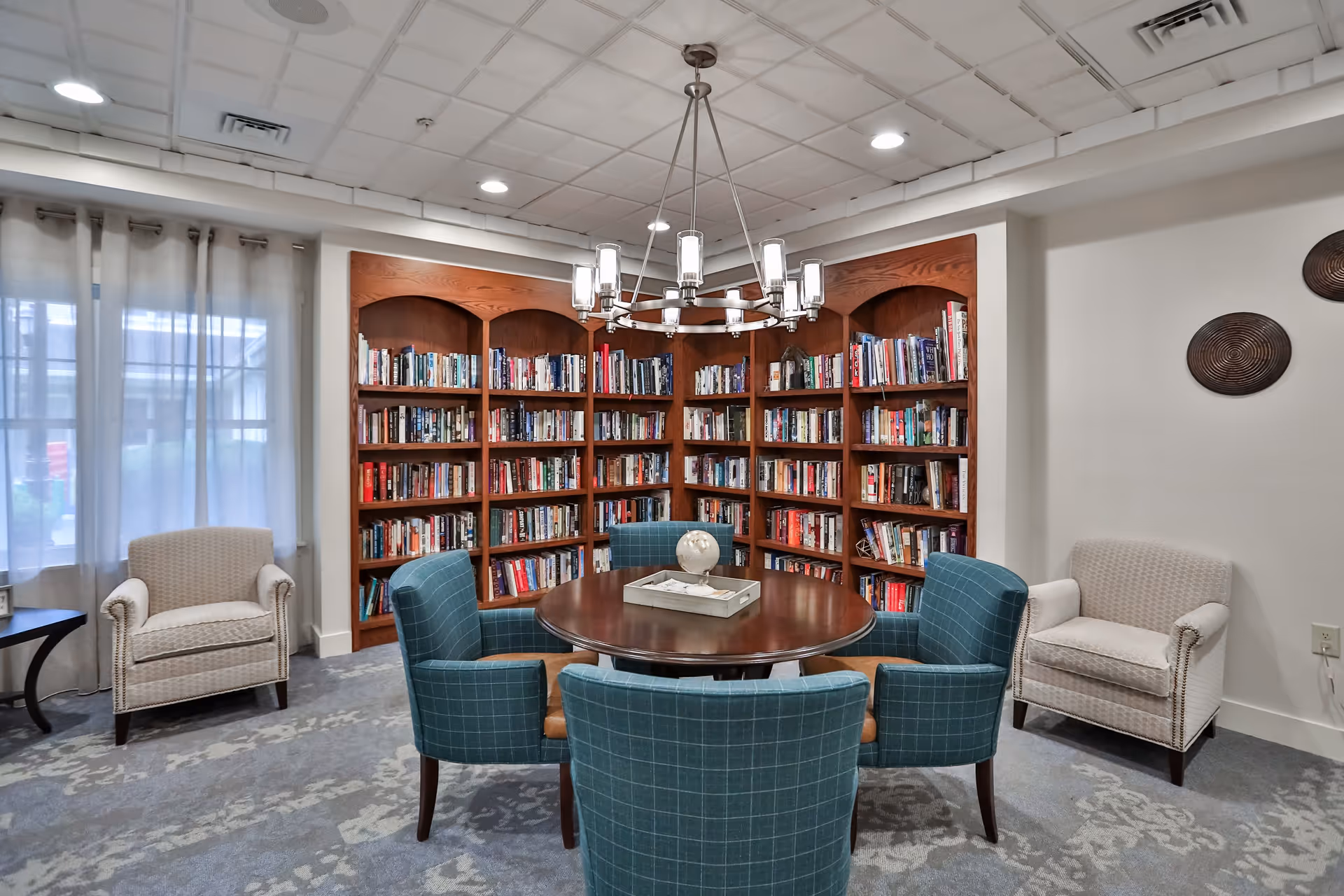A cozy reading room with a round wooden table surrounded by four blue upholstered chairs. Behind the table is a large wooden bookshelf filled with books. Two beige armchairs are placed near the windows with sheer curtains on the left and on the right wall are two decorative circular wall hangings. A modern chandelier hangs from the ceiling above the table.
