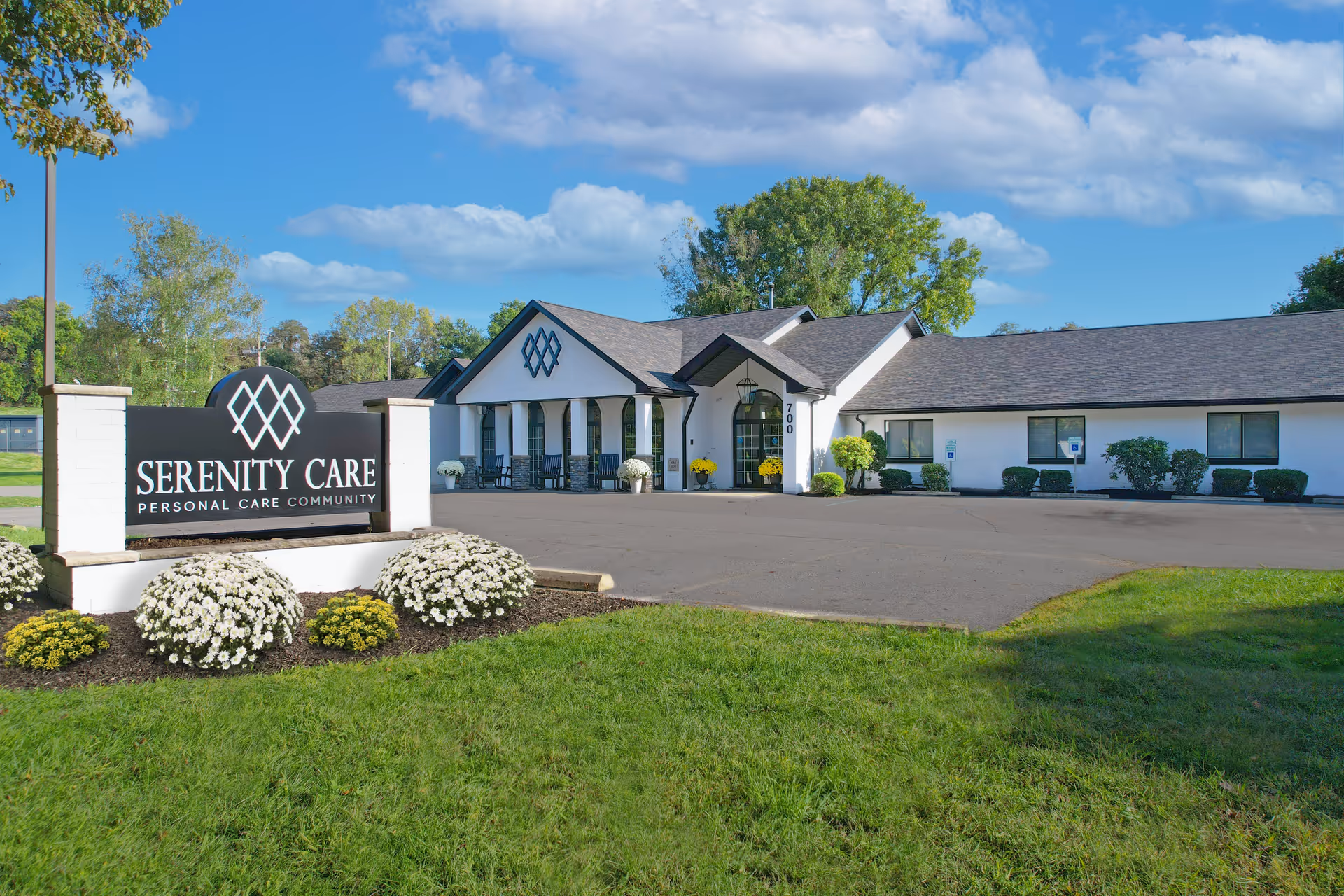 Exterior view of Serenity Care Personal Care Community building with a large sign in front, surrounded by green grass, white and yellow flowers, and trees under a blue sky with some clouds.