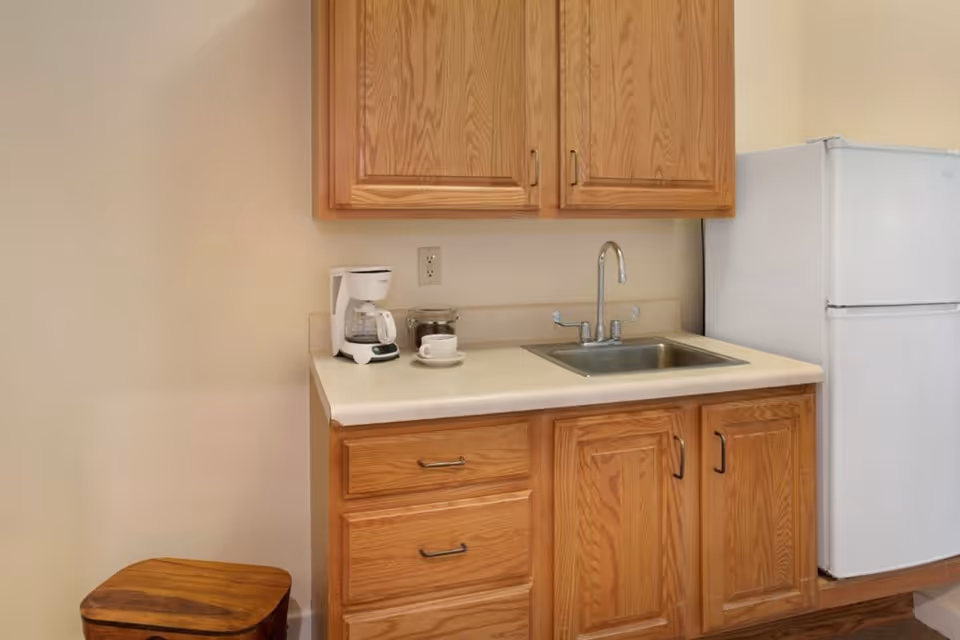 A small kitchenette area with wooden cabinets, a white countertop, a stainless steel sink with a faucet, a white coffee maker with a cup and saucer, and a white refrigerator. There is also a wooden stool partially visible on the left side.