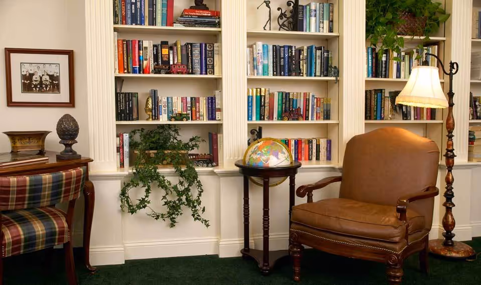 Cozy reading nook with built-in bookshelves filled with books, a brown leather armchair, a globe on a wooden stand, a floor lamp with a fringed shade, a plaid upholstered chair, a wooden table, and a framed black and white photograph on the wall.