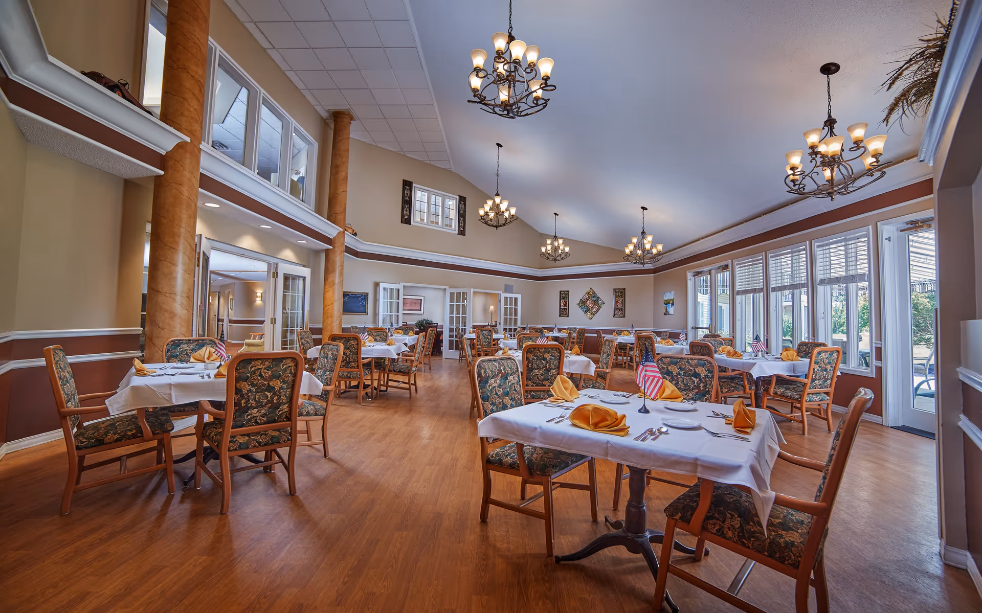 A spacious dining room in a senior living facility with multiple tables covered in white tablecloths, each set with plates, silverware, and folded yellow napkins. The chairs have floral upholstery, and small American flags are placed on some tables. The room features wooden floors, tall decorative columns, large windows letting in natural light, and elegant chandeliers hanging from a high ceiling.