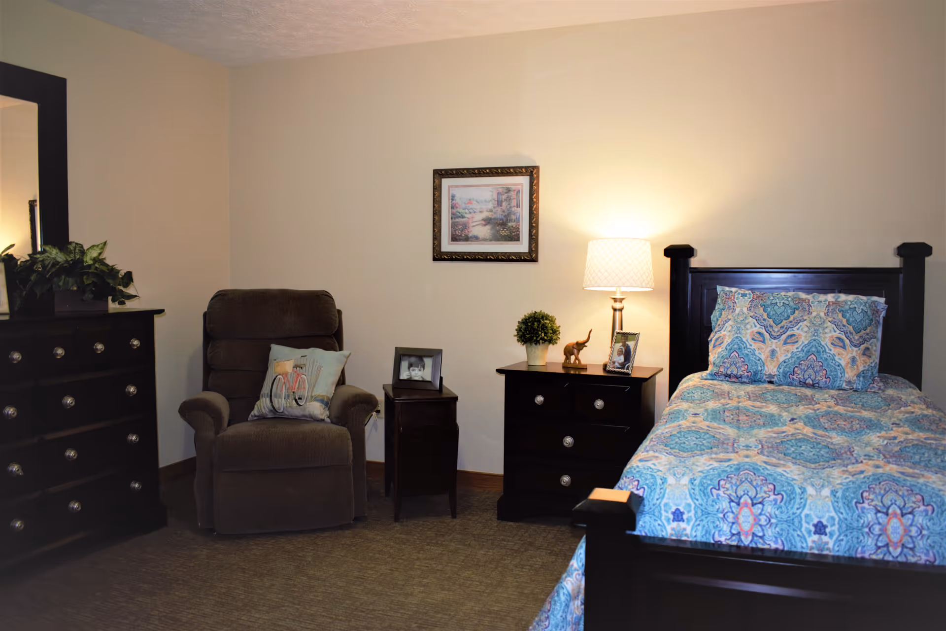 A cozy bedroom in an assisted living facility featuring a single bed with a colorful patterned bedspread and matching pillows. Next to the bed is a dark wooden nightstand with a table lamp, a small potted plant, a framed photo, and a small elephant figurine. To the left is a comfortable brown armchair with a decorative pillow, a small side table with a framed photo, and a dark wooden dresser with a large mirror and a plant on top. A framed picture hangs on the beige wall above the nightstand.