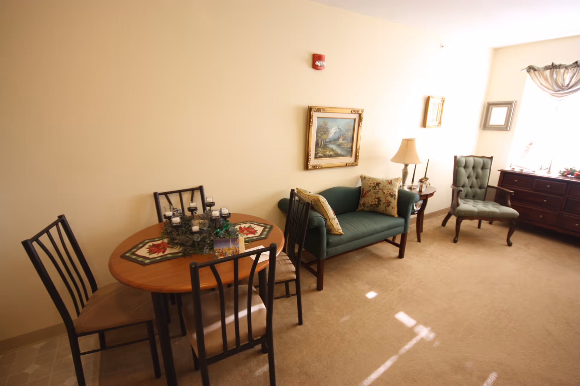 A cozy living area with a small wooden dining table surrounded by four chairs, a green upholstered loveseat with floral cushions, a wooden side table with a lamp, a green cushioned armchair, and a wooden dresser near a window with sheer curtains. The walls are decorated with framed paintings.