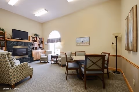 A cozy living room area with beige walls and carpeted floor. The room features a fireplace with a black marble surround, a flat-screen TV above it, and built-in wooden shelves filled with books and plants. There are several upholstered armchairs arranged around a small wooden table near a window with an arched top and a valance. A dining table with six chairs is positioned on the right side of the room, accompanied by a floor lamp and wall art.