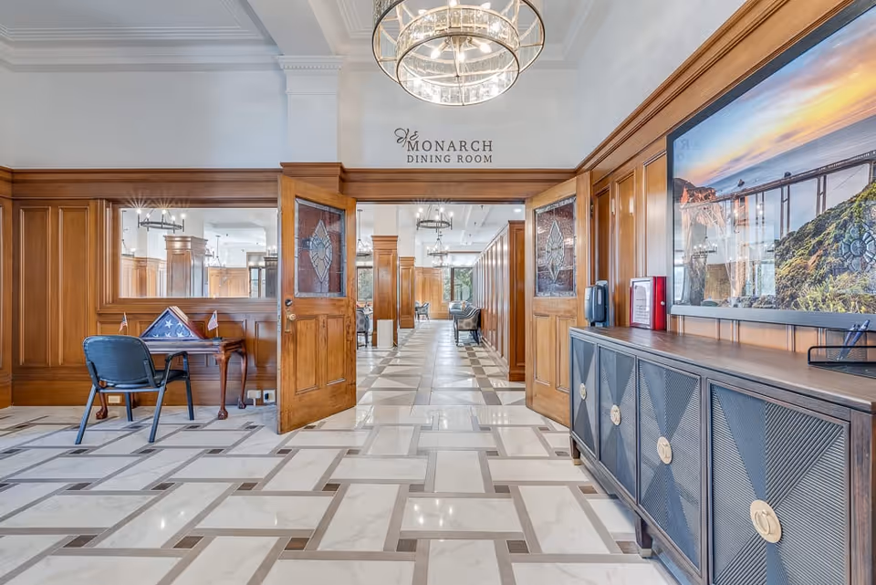 Interior view of the Monarch Dining Room at Pacific Grove Senior Living, featuring an open wooden double door with stained glass panels, wood-paneled walls, a tiled floor with a geometric pattern, a chandelier hanging from the ceiling, a small table with a folded American flag and two small flags on the left, and a sideboard with a large framed photograph of a bridge on the right.