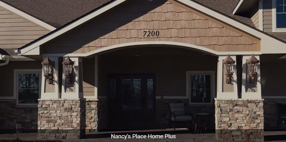 Entrance of a residential-style building with stone pillars, an arched porch, the number 7200 above the doorway, and outdoor chairs on the stoop.