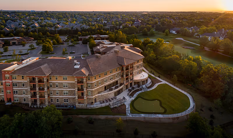 Aerial view of Fairways of Ironhorse Independent Living facility at sunset, showing a large multi-story building with balconies, surrounded by green lawns, trees, and a putting green. Residential neighborhood and parking lot are visible in the background.