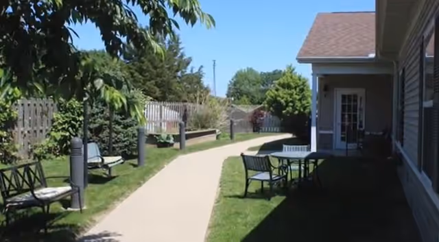 Sunny outdoor courtyard walkway with benches, a table and chairs, landscaping, and a building entrance.