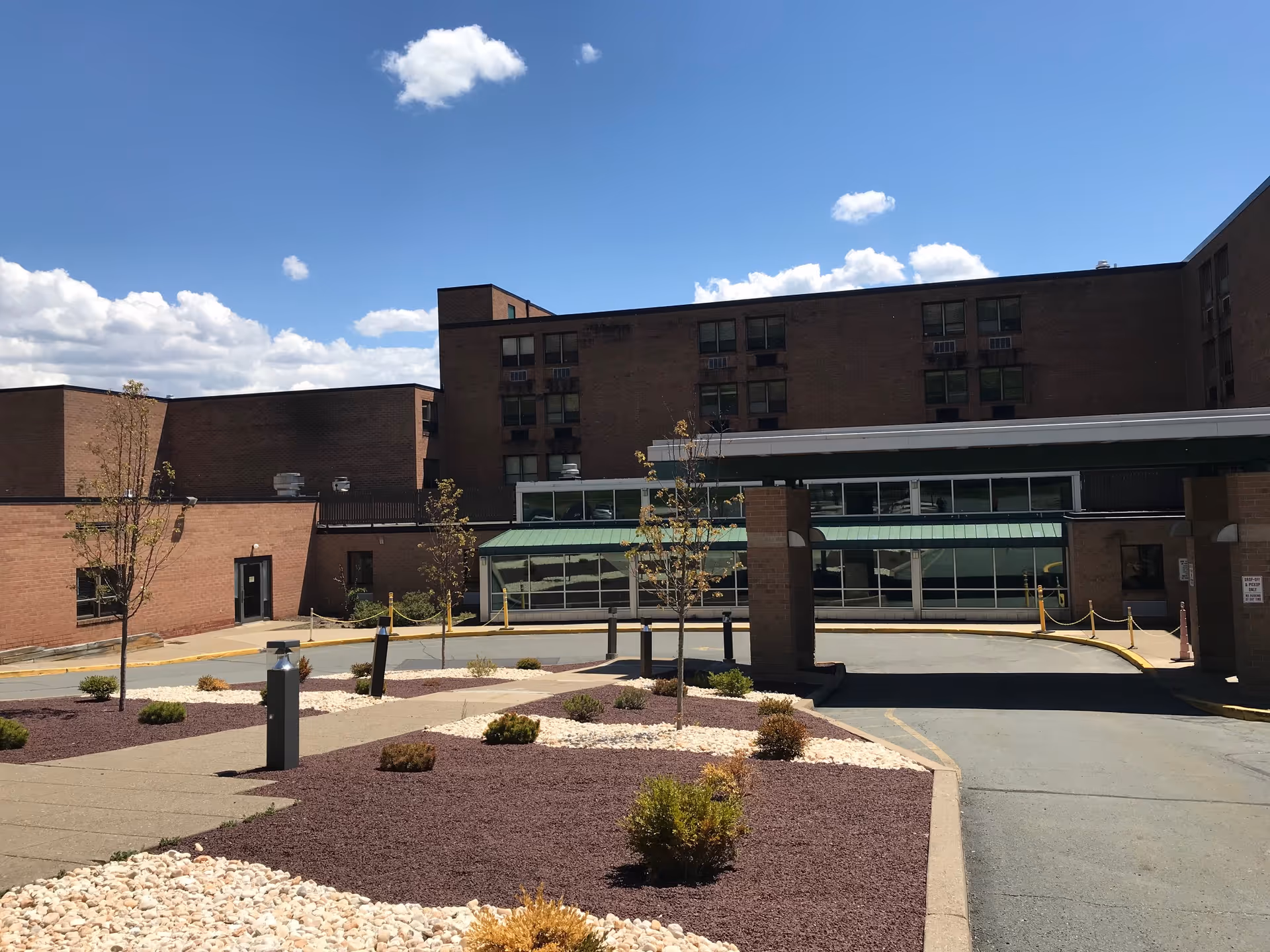 Exterior view of Allied Services Skilled Nursing & Rehabilitation Center showing a multi-story brick building with several windows, a covered entrance driveway, small landscaped areas with young trees and shrubs, and a clear blue sky with a few clouds.