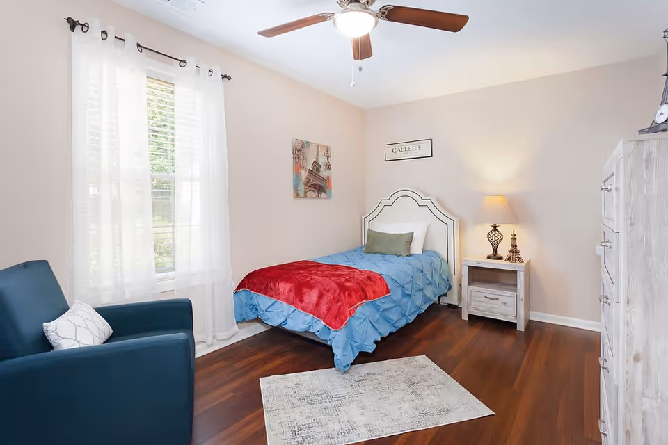 A cozy bedroom with a single bed covered in a blue comforter and a red throw blanket. There is a green pillow and a white pillow on the bed. Next to the bed is a white nightstand with a lamp and a small Eiffel Tower decoration. A blue armchair with a white pillow is positioned near a window with sheer white curtains. The room has wooden flooring, a ceiling fan with a light, and light-colored walls with two framed pictures hanging above the bed.