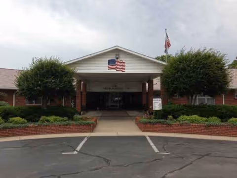 Front entrance of a brick senior living facility with flags and landscaped shrubs.