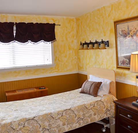 A single bed with floral patterned bedding and a brown pillow in a room with yellow textured walls and a wooden wainscot. There is a window with white blinds and a dark brown valance, a wooden nightstand with a lamp, and a framed floral picture on the wall.