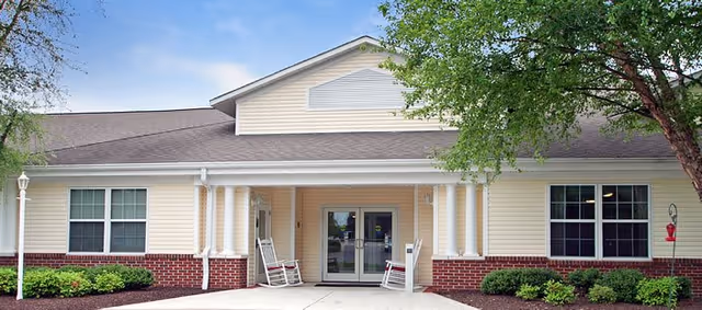 Front exterior view of a single-story senior living facility building with beige siding and red brick accents. The entrance has double glass doors with two white rocking chairs on the porch. There are windows on either side of the entrance and small bushes along the building. A tree is visible on the right side under a partly cloudy sky.