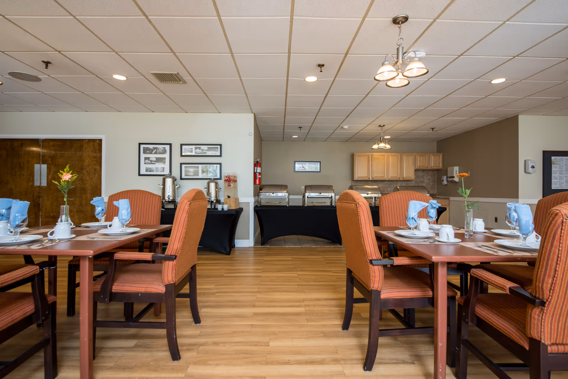 Dining room in a senior living facility with wooden tables set with white cups, plates, and blue napkins. Orange upholstered chairs surround the tables. In the background, there are buffet tables with chafing dishes and coffee dispensers, light wood cabinets, and framed pictures on the walls. The room has a light-colored ceiling with recessed lighting and hanging light fixtures.