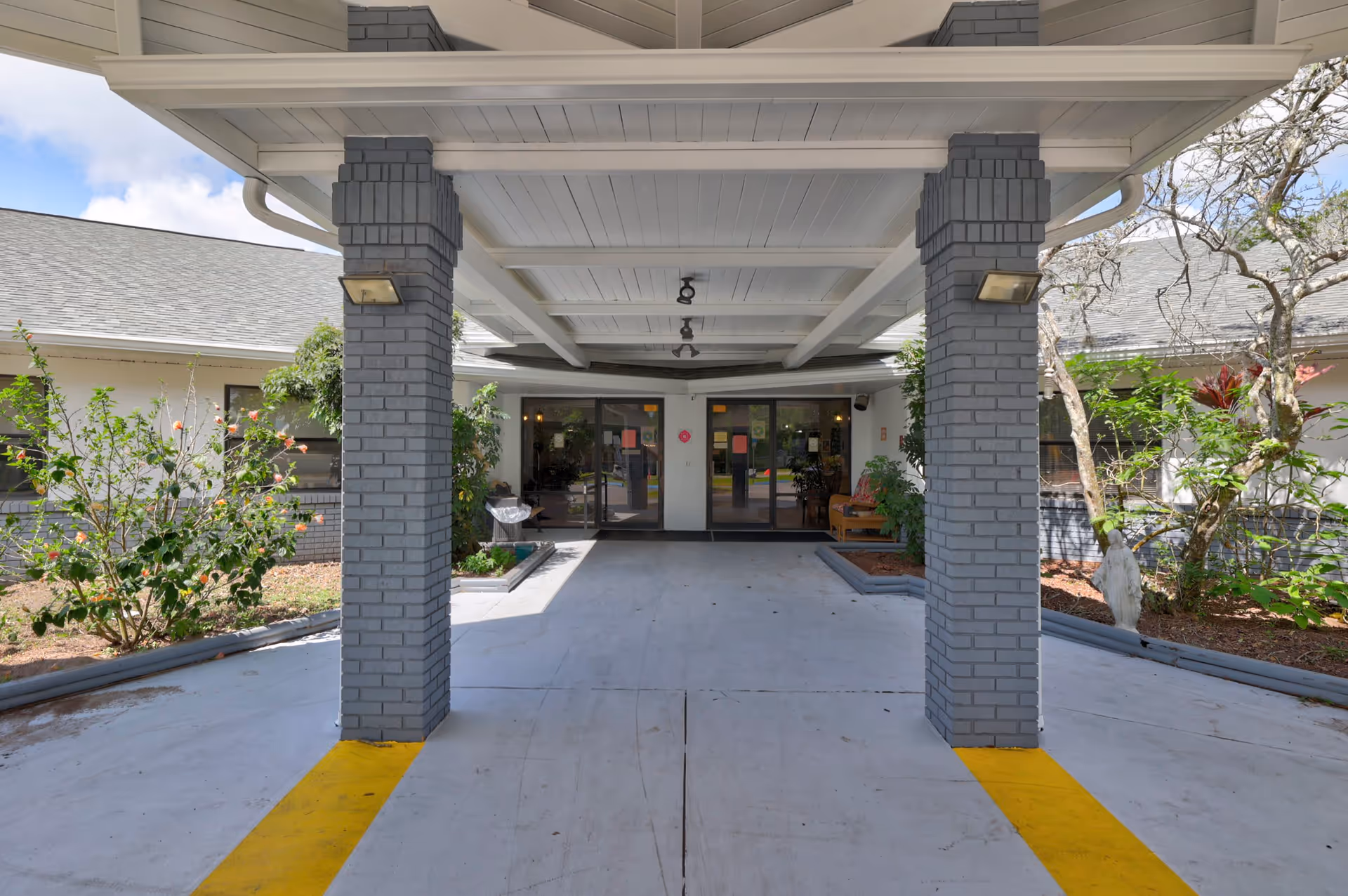 Covered entrance area of a building with two gray brick pillars and a white ceiling. There are glass double doors at the end of the driveway, flanked by plants and shrubs on either side. The driveway has yellow lines painted near the pillars, and the sky is partly cloudy.