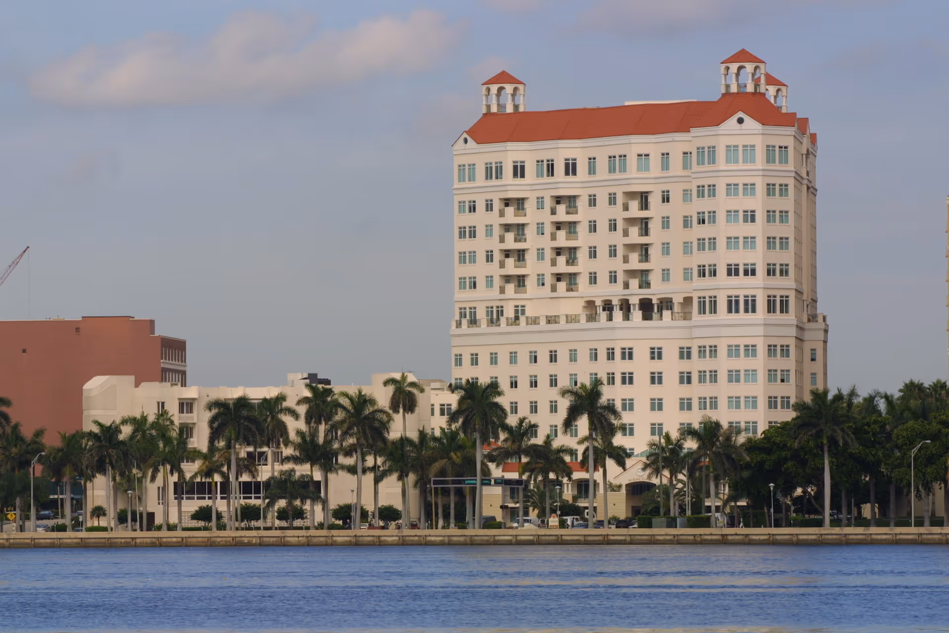 A multi-story building with a red roof and multiple windows, situated behind a row of palm trees along a waterfront under a partly cloudy sky.