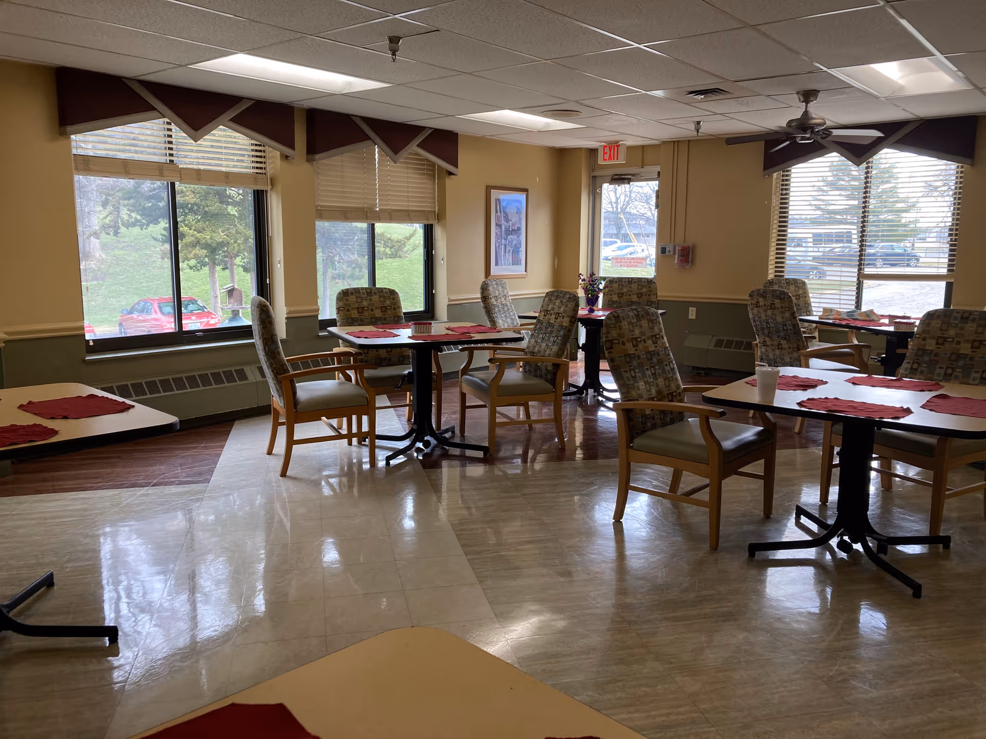 A dining room in an assisted living facility with several tables and cushioned chairs arranged neatly. Each table has red placemats and some have small items like a cup or a flower vase. Large windows allow natural light to brighten the room, and outside greenery and parked cars are visible through the windows.