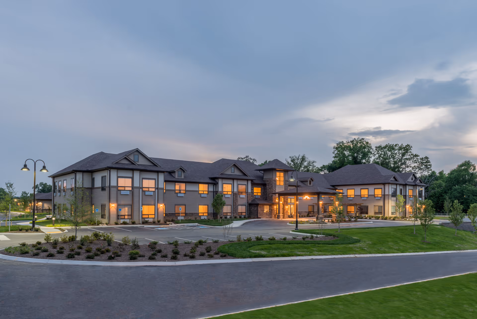 Exterior view of The Pointe at Lifespring Senior Living building at dusk with lights glowing from windows, surrounded by landscaped greenery and a paved driveway.