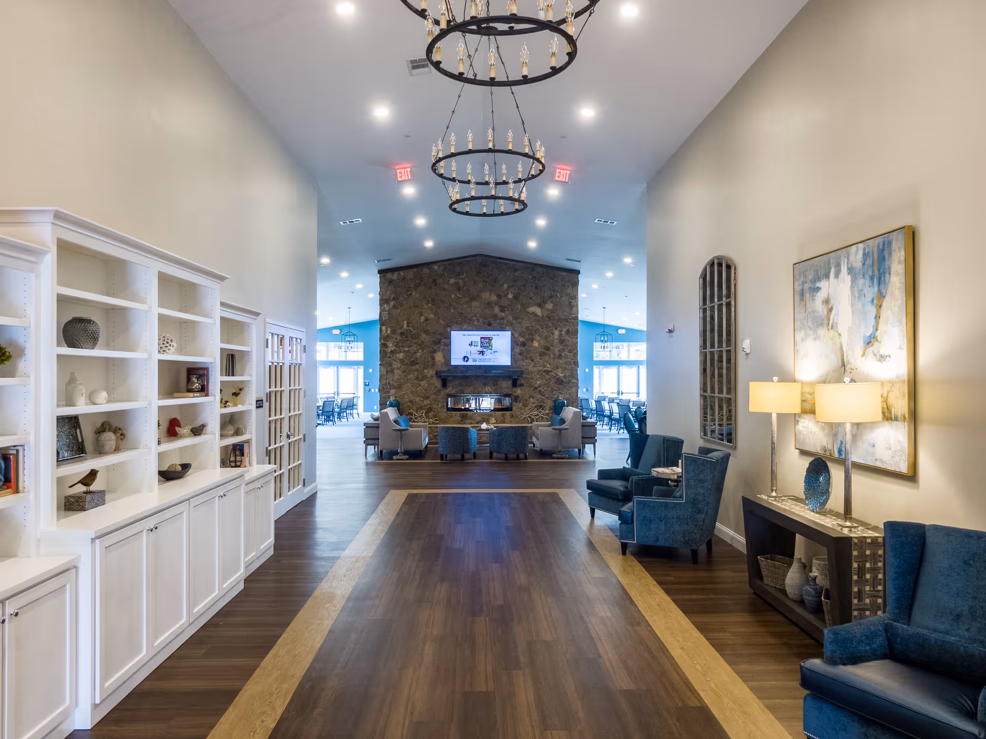 A spacious and well-lit interior hallway leading to a seating area with a stone fireplace and a mounted TV. The hallway features wooden flooring with a lighter border, white built-in shelves on the left, and blue upholstered chairs and a console table with lamps and decorative items on the right. Two large circular chandeliers hang from the ceiling.