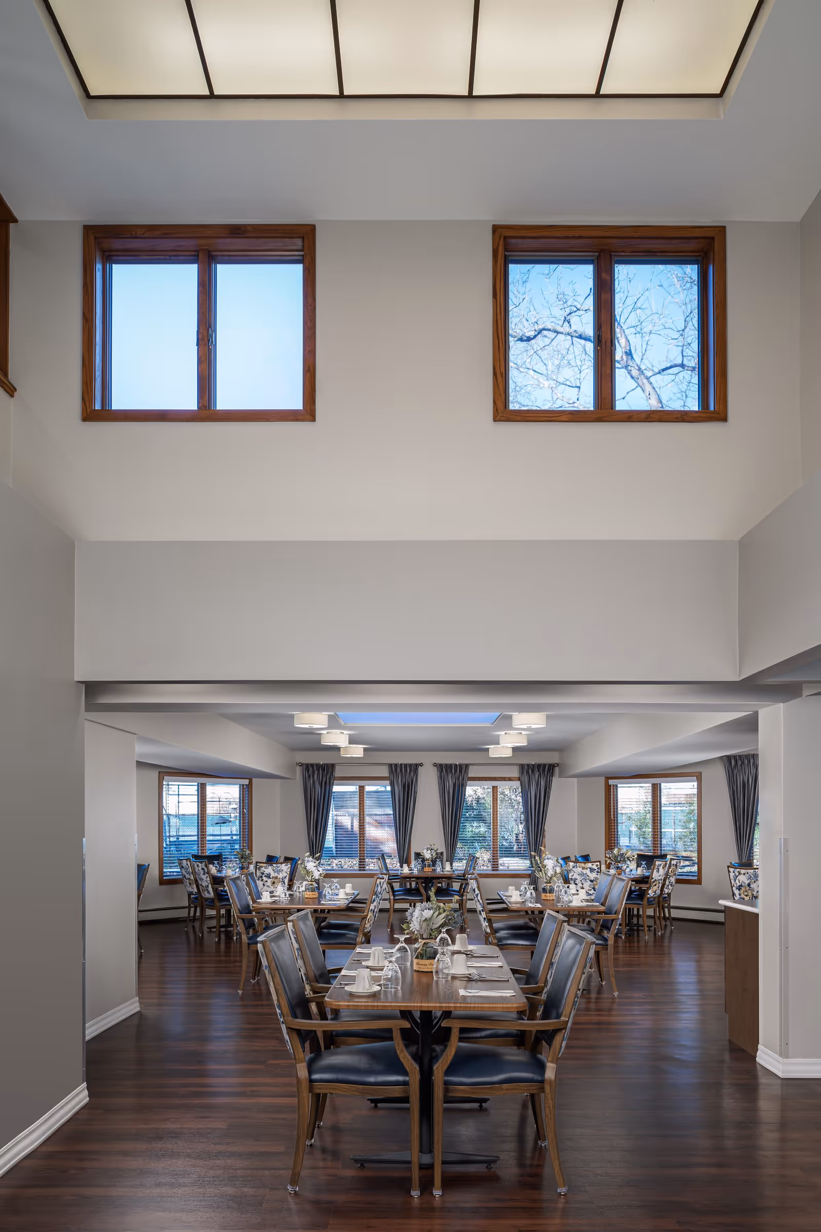 Dining room with rows of set wooden tables and chairs, large windows, and high ceilings with overhead lighting.