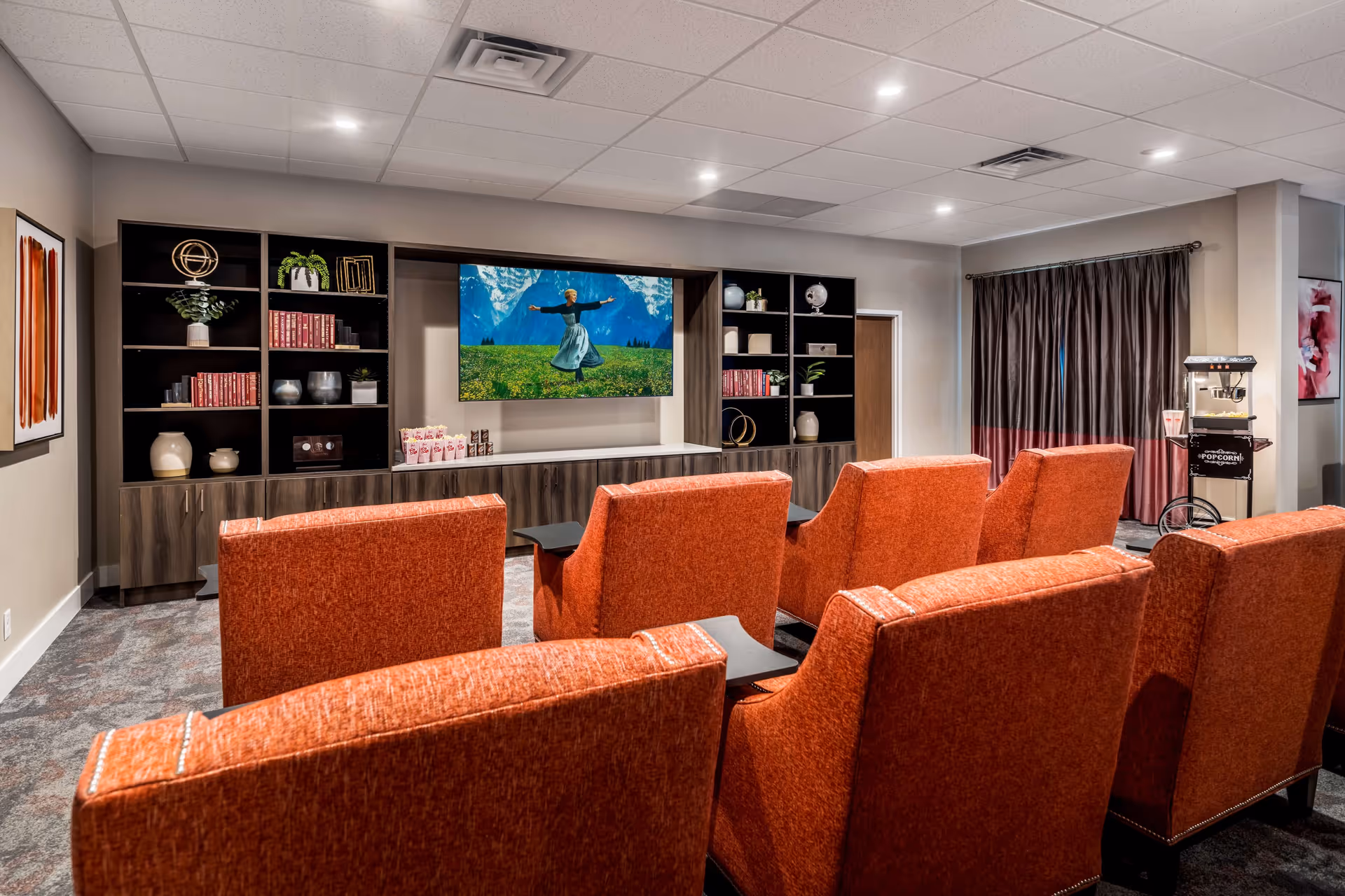 Small theater-style common room with rows of orange upholstered seats facing a wall-mounted TV and shelving, with a popcorn machine at the side.