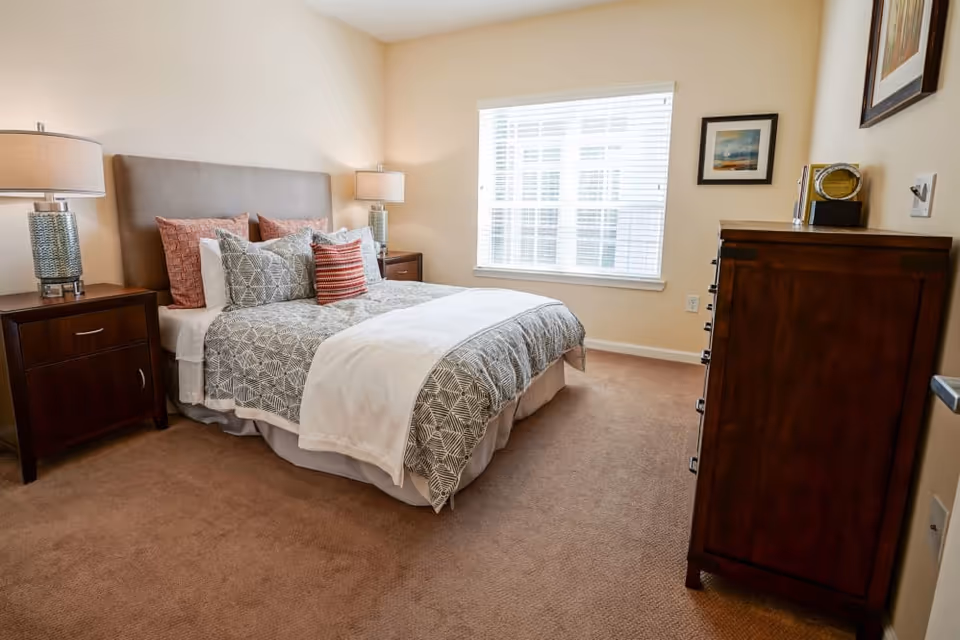 A cozy bedroom featuring a neatly made bed with decorative pillows, two bedside lamps, and a wooden dresser. Natural light streams in through a window with blinds, illuminating the warm color palette of the room.