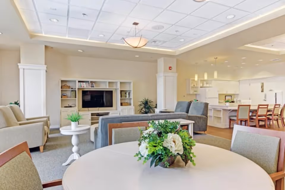 A bright and spacious senior living common area featuring a round table with a floral centerpiece in the foreground, comfortable armchairs and sofas arranged around a TV on a white entertainment unit, and a kitchen and dining area with wooden chairs and pendant lighting in the background.
