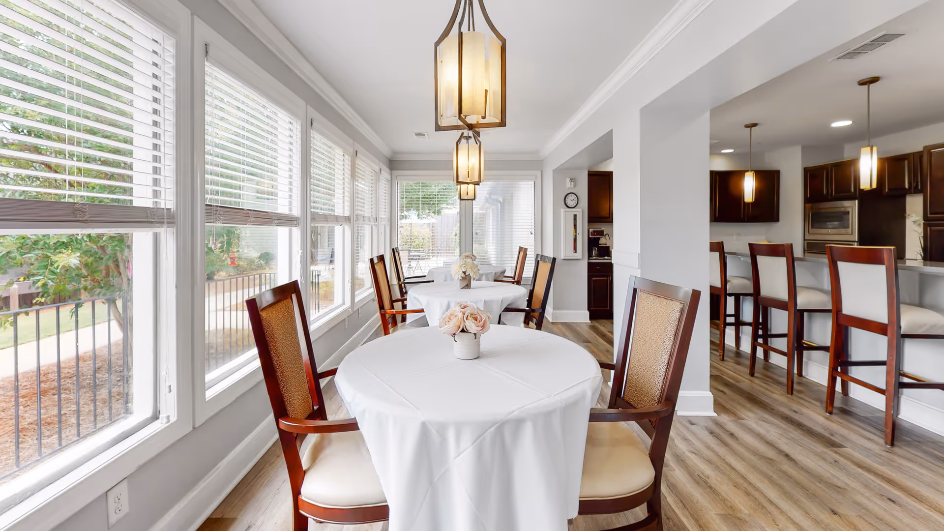 A bright dining area with round tables covered in white tablecloths, each adorned with a small vase of flowers. Wooden chairs with cushioned seats surround the tables. Large windows with white blinds line one side, letting in natural light and offering a view of the outdoor greenery. Pendant lights hang from the ceiling, and a kitchen area with a breakfast bar and high chairs is visible in the background.