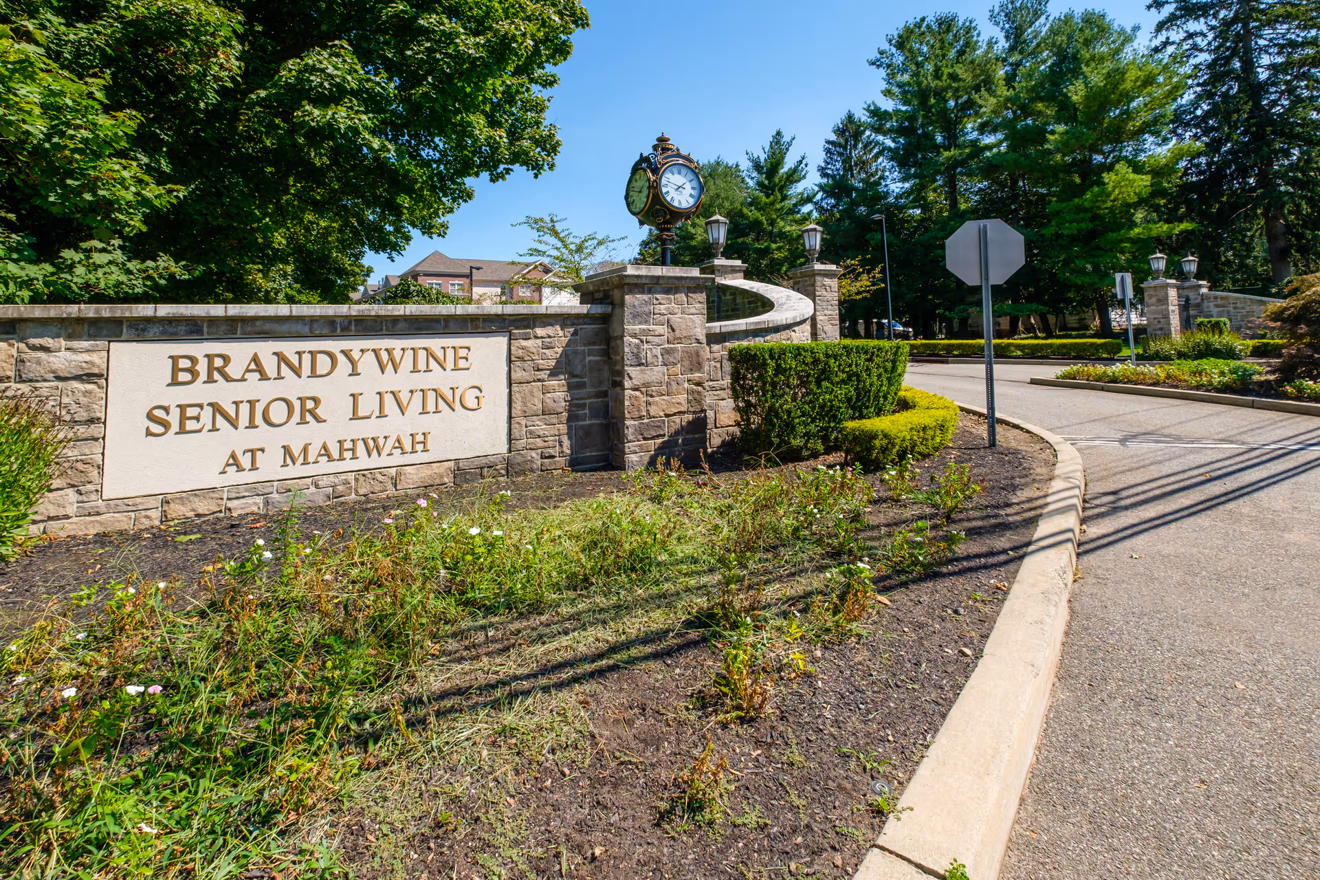 Entrance to Brandywine Senior Living at Mahwah featuring a stone wall with the facility's name engraved, surrounded by greenery and a decorative clock mounted on a stone pillar under a clear blue sky.