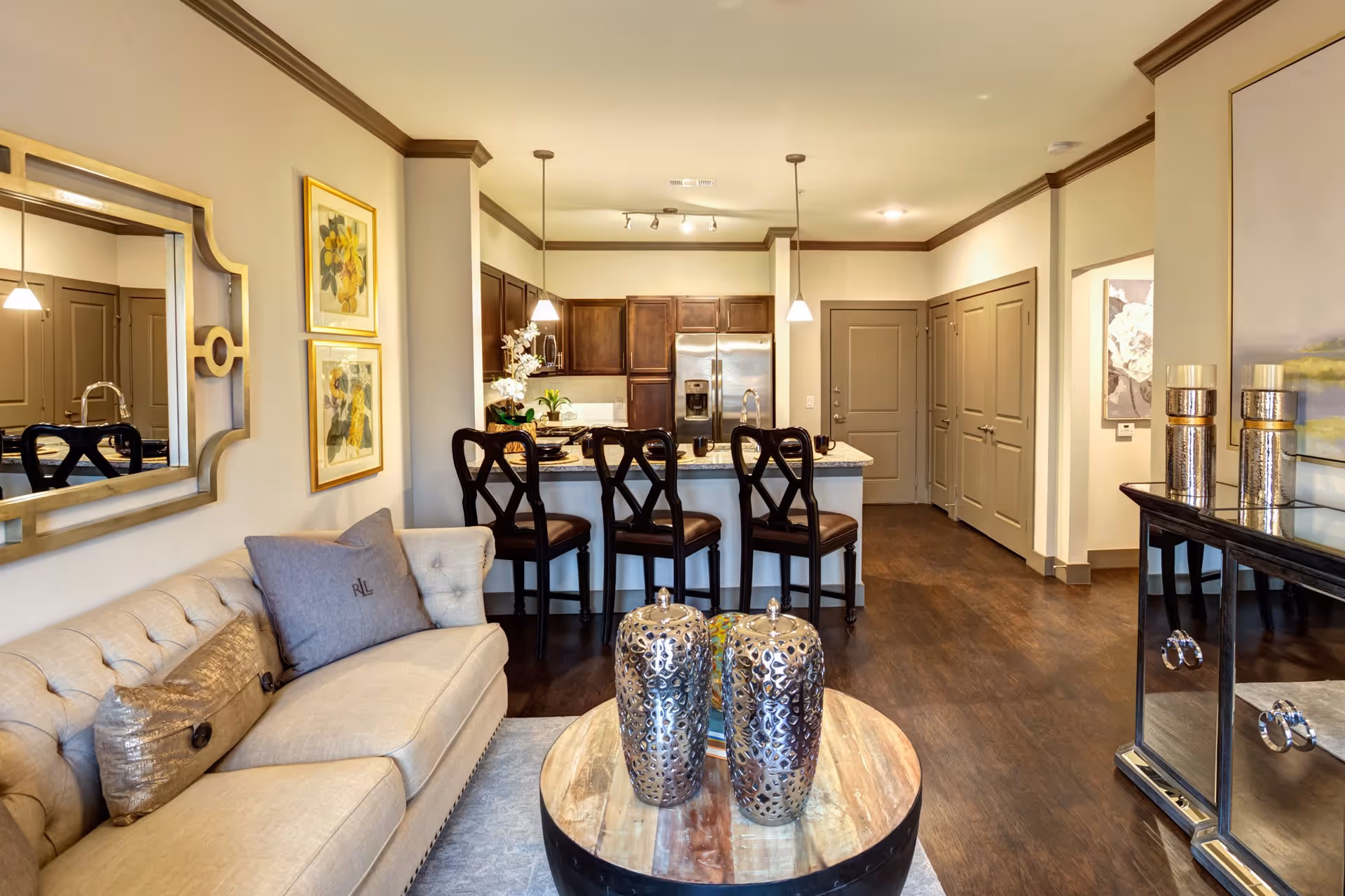 Open-concept living area with a beige tufted sofa, round wood coffee table topped with decorative vases, and a kitchen island with three barstools.