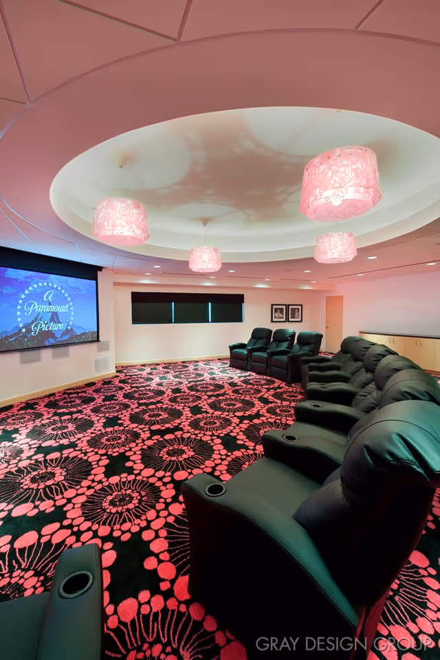 A home theater room with black leather reclining chairs arranged in rows facing a large screen displaying 'A Paramount Picture'. The room features a patterned carpet with pink and black floral designs, pink pendant lights hanging from a circular recessed ceiling, and a window with black blinds.