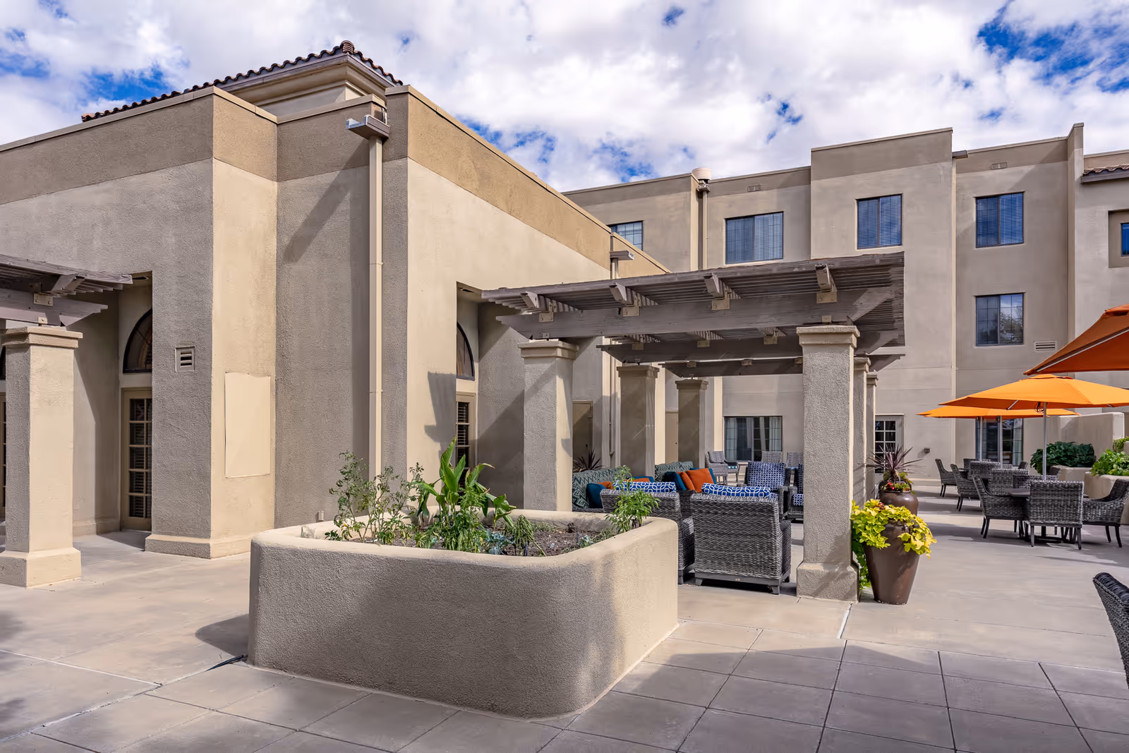 Outdoor patio area at Fairwinds - Rio Rancho featuring a covered seating area with wicker chairs and colorful cushions, potted plants, and orange umbrellas under a partly cloudy sky.