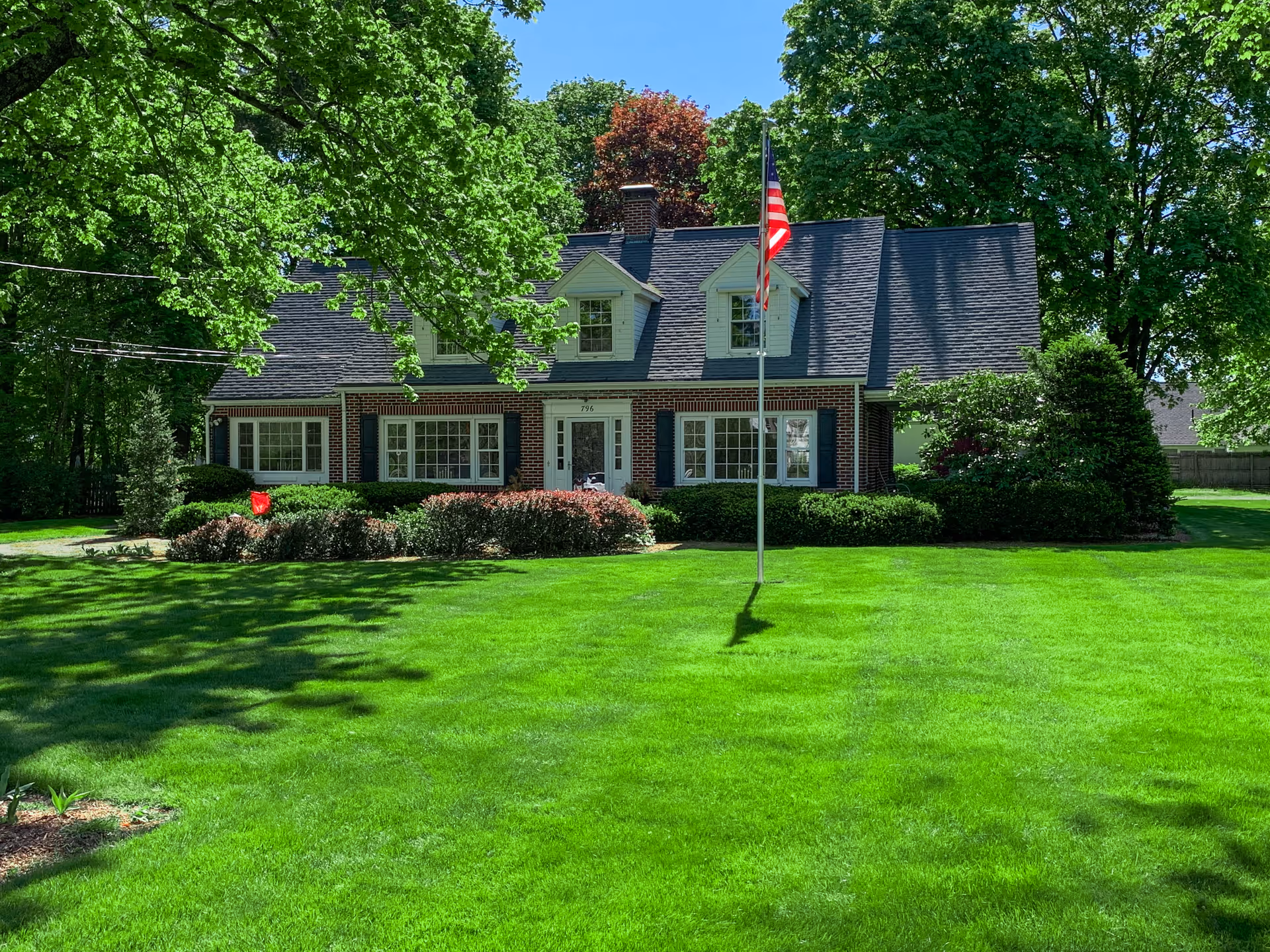 A brick residential building with a dark shingled roof and white-framed windows, surrounded by lush green trees and a well-maintained lawn. An American flag is mounted on a flagpole in front of the building.