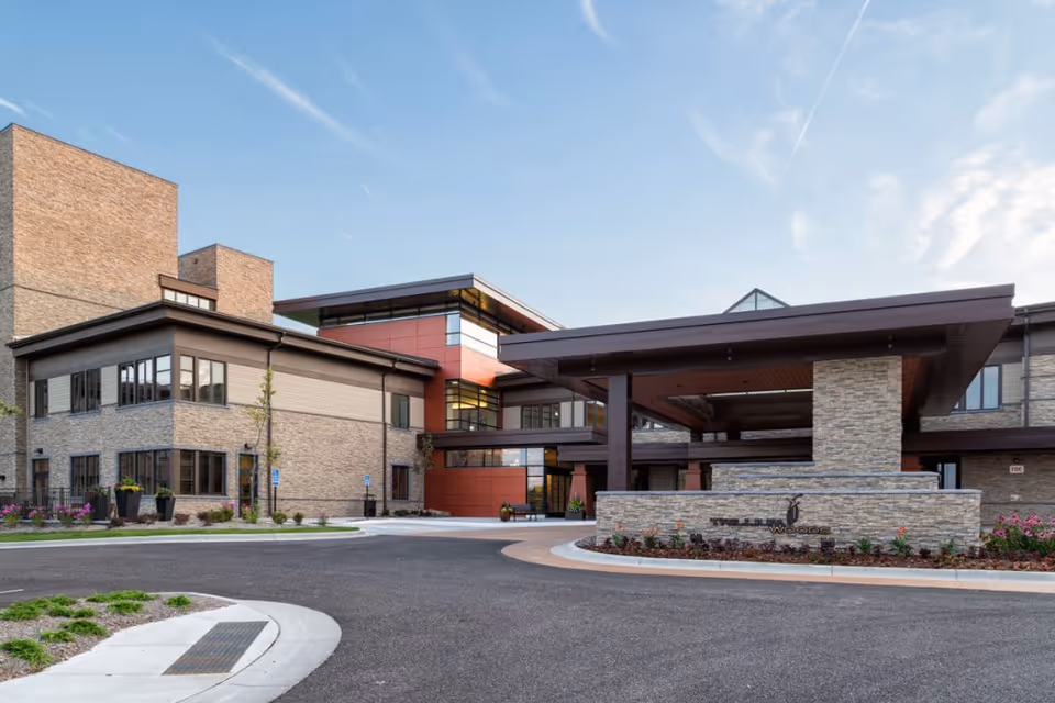 Exterior view of a modern senior living facility building named Trillium Woods, featuring a covered entrance with stone and brick facade, large windows, and landscaped surroundings under a clear sky.