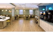 A spacious dining area with multiple tables and chairs arranged neatly. The room has light wood flooring, greenish walls, and several windows letting in natural light. On the right side, there is a counter with coffee machines and beverage dispensers. The ceiling has recessed lighting and two hanging pendant lights above the beverage area.