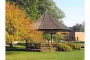 A wooden gazebo on a grassy lawn surrounded by trees and shrubs under a clear sky.