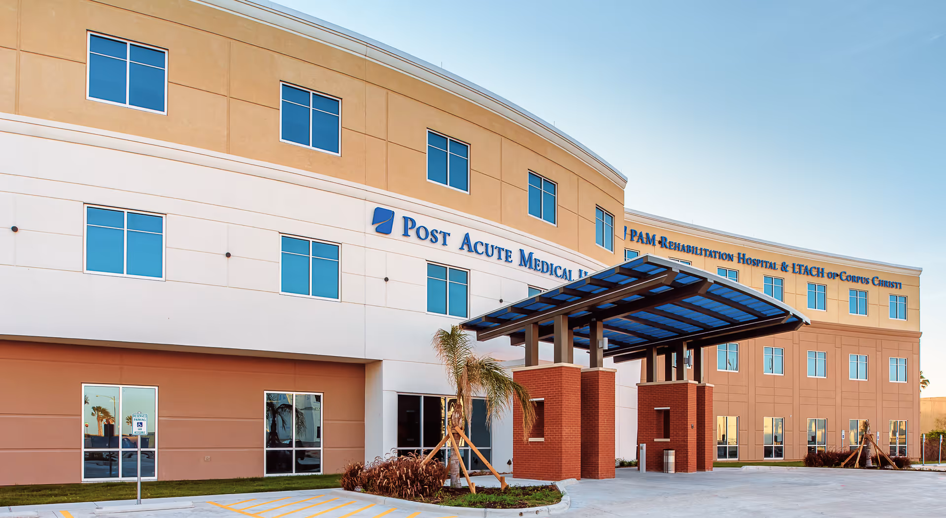 Exterior view of PAM Health Rehabilitation Hospital of Corpus Christi, a modern three-story building with large windows, a covered entrance with brick pillars, and a clear blue sky.