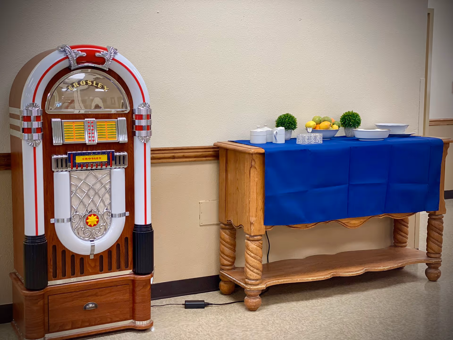 A vintage Crosley jukebox stands next to a wooden table covered with a blue cloth. On the table are two small green potted plants, a bowl of assorted fruit, white ceramic containers, and a decorative glass box. The setting appears to be indoors with beige walls and a tiled floor.