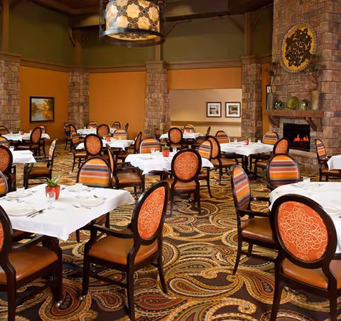 A spacious dining room with multiple tables covered in white tablecloths, each set with plates, glasses, and silverware. The chairs have wooden frames with orange and striped upholstery. The room features a patterned carpet, stone pillars, a fireplace with a decorative piece above it, and warm lighting from a large hanging lamp.