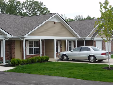 Single-story senior living building front with a covered porch, a parked silver sedan, and a green lawn.
