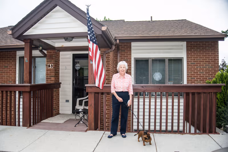 An elderly woman standing outside a brick and white siding building with a wooden porch railing, holding a small dog on a leash. An American flag is mounted on the porch near the entrance door.