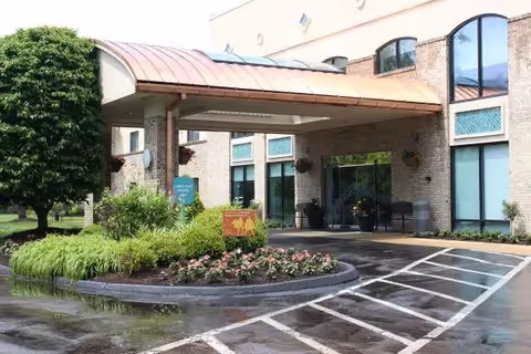 Front entrance of a senior living facility with a covered drop-off canopy, landscaped roundabout, and glass entry doors.
