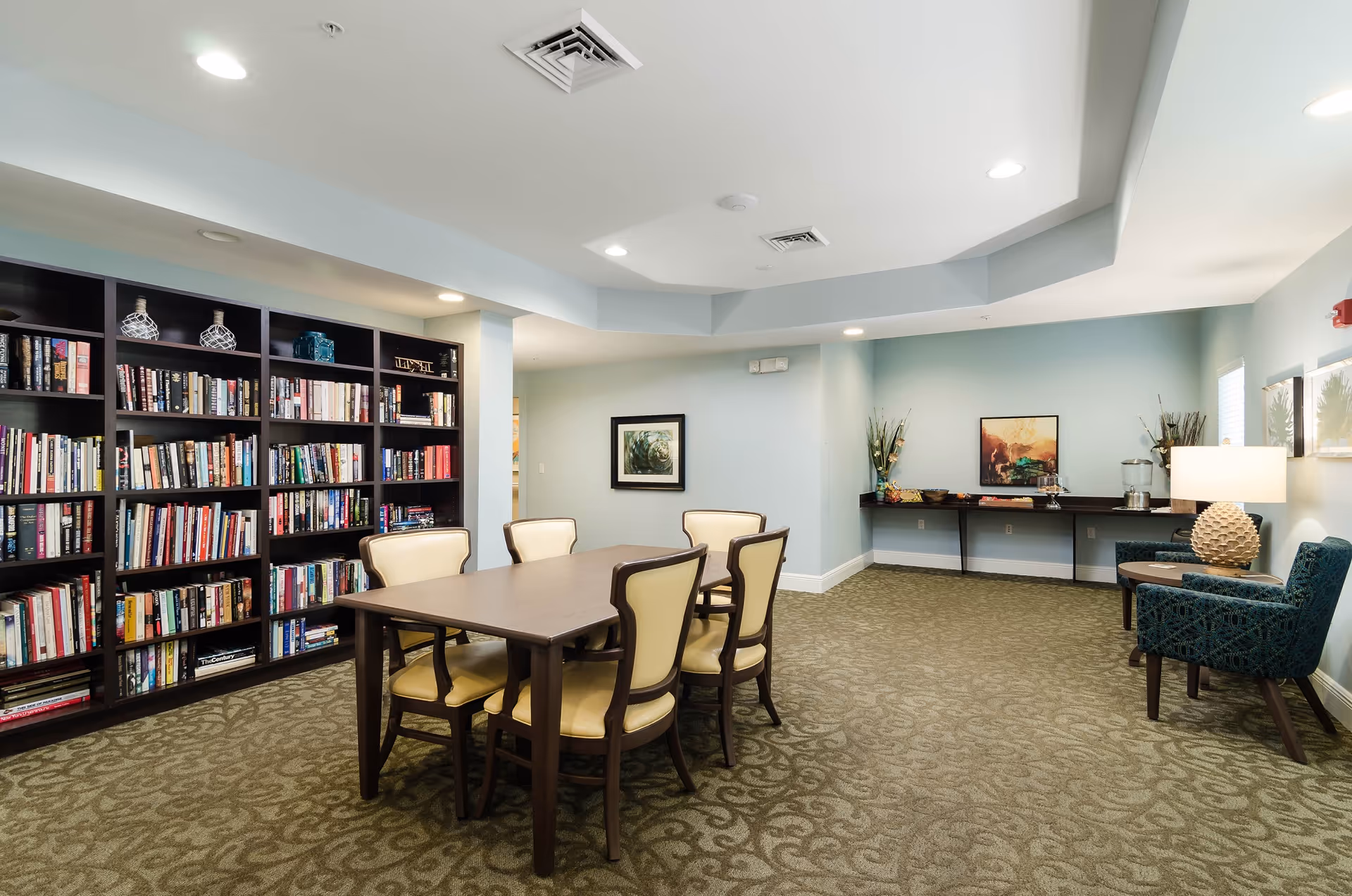 A bright communal lounge with a central table and chairs, a large bookshelf, and a seating area with artwork and a lamp.