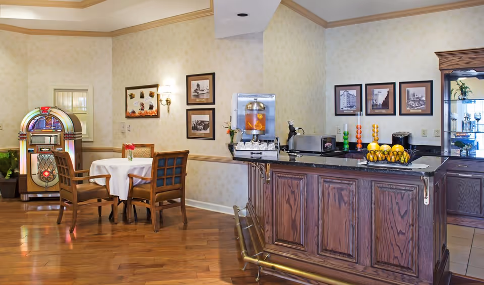 A cozy interior space featuring a small round table with four wooden chairs, a vintage jukebox, and a wooden counter with a black countertop. The counter has a beverage dispenser, a toaster, and a basket of fruit. The walls are decorated with framed black and white photographs and a small wall lamp.