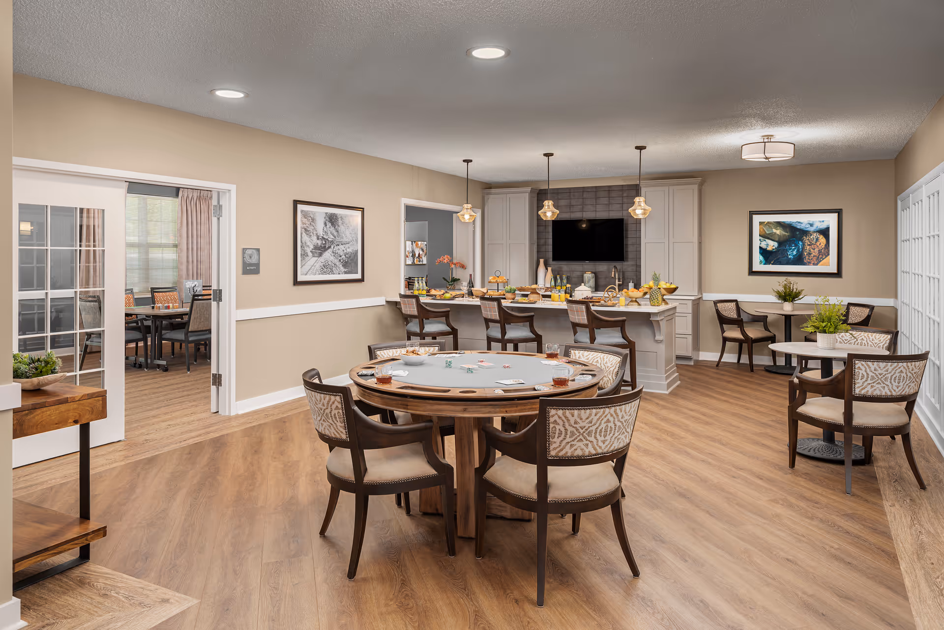 A spacious and well-lit common area in a senior living facility featuring a round table with six chairs set up for a card game, a kitchen island with bar stools and pendant lights, and additional small tables with chairs along the walls. The room has wood flooring, beige walls, framed artwork, and a flat-screen TV mounted above the kitchen counter.