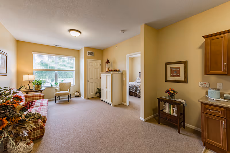 A cozy living room area in a senior living facility with beige walls and carpet. The room features a plaid sofa with floral cushions, a small table with a lamp and plant near a large window with blinds, a beige armchair, a white cabinet, and a small wooden side table with books and a flower vase. A doorway leads to a bedroom with a bed visible. Part of a kitchen counter and cabinets are seen on the right side.
