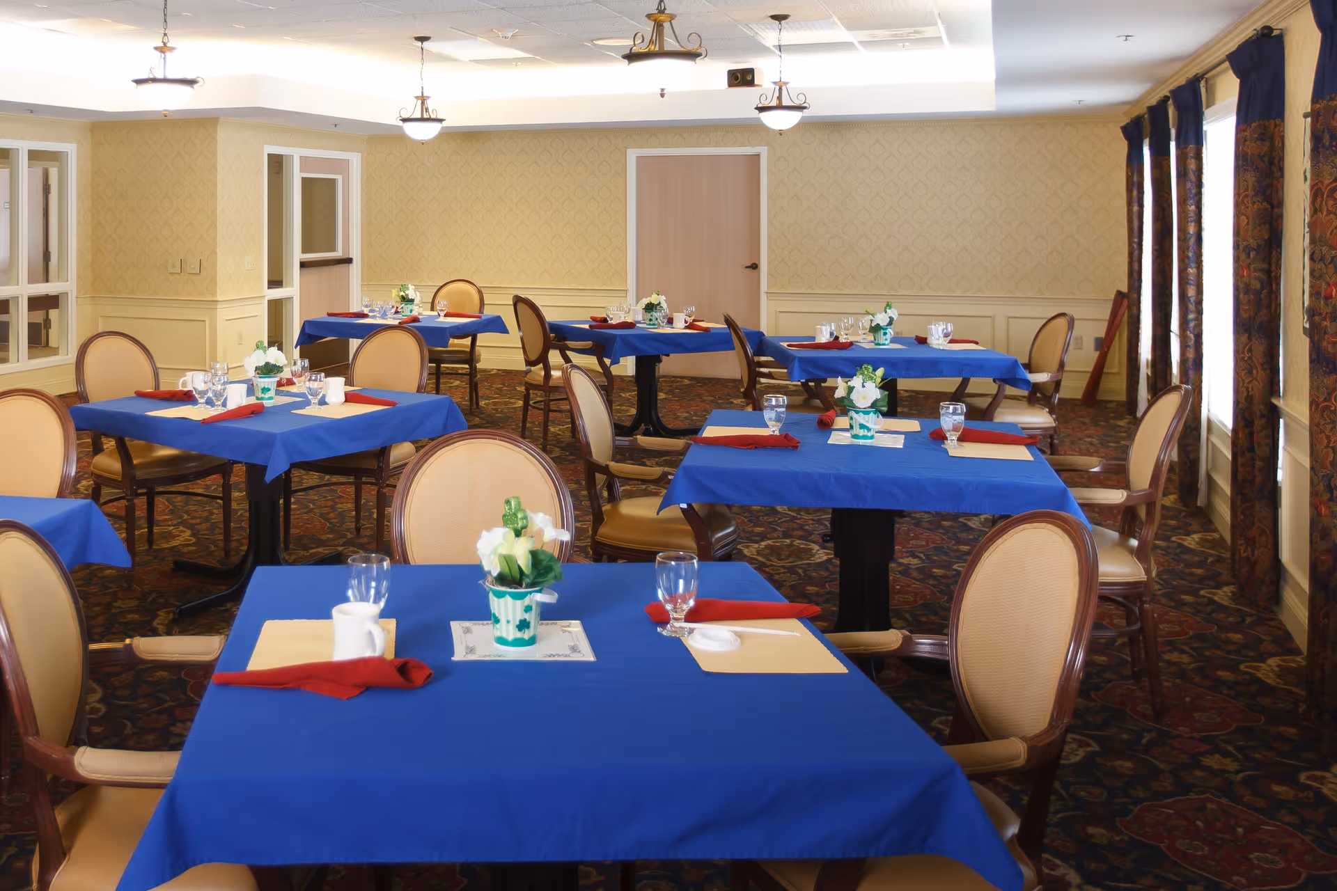 A dining room with several tables draped in blue tablecloths, set with place settings and surrounded by upholstered chairs.