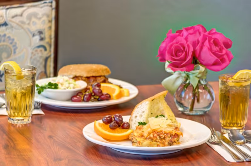 A dining table set with two plates of food, each accompanied by a glass of iced tea with a lemon wedge. One plate has a sandwich, coleslaw, grapes, and orange slices. The other plate has a slice of lasagna, a piece of bread, grapes, and orange slices. A small vase with bright pink roses tied with a green ribbon is also on the table.