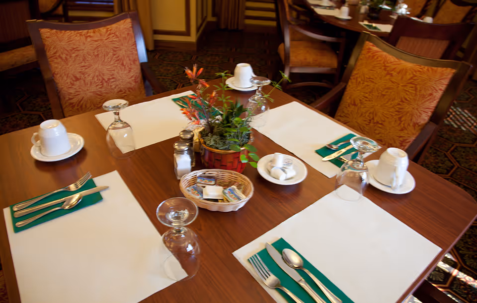 A dining table set for four with white placemats, green napkins, silverware, upside-down wine glasses, white cups on saucers, a small basket with butter packets, salt and pepper shakers, and a small plant centerpiece in a basket.