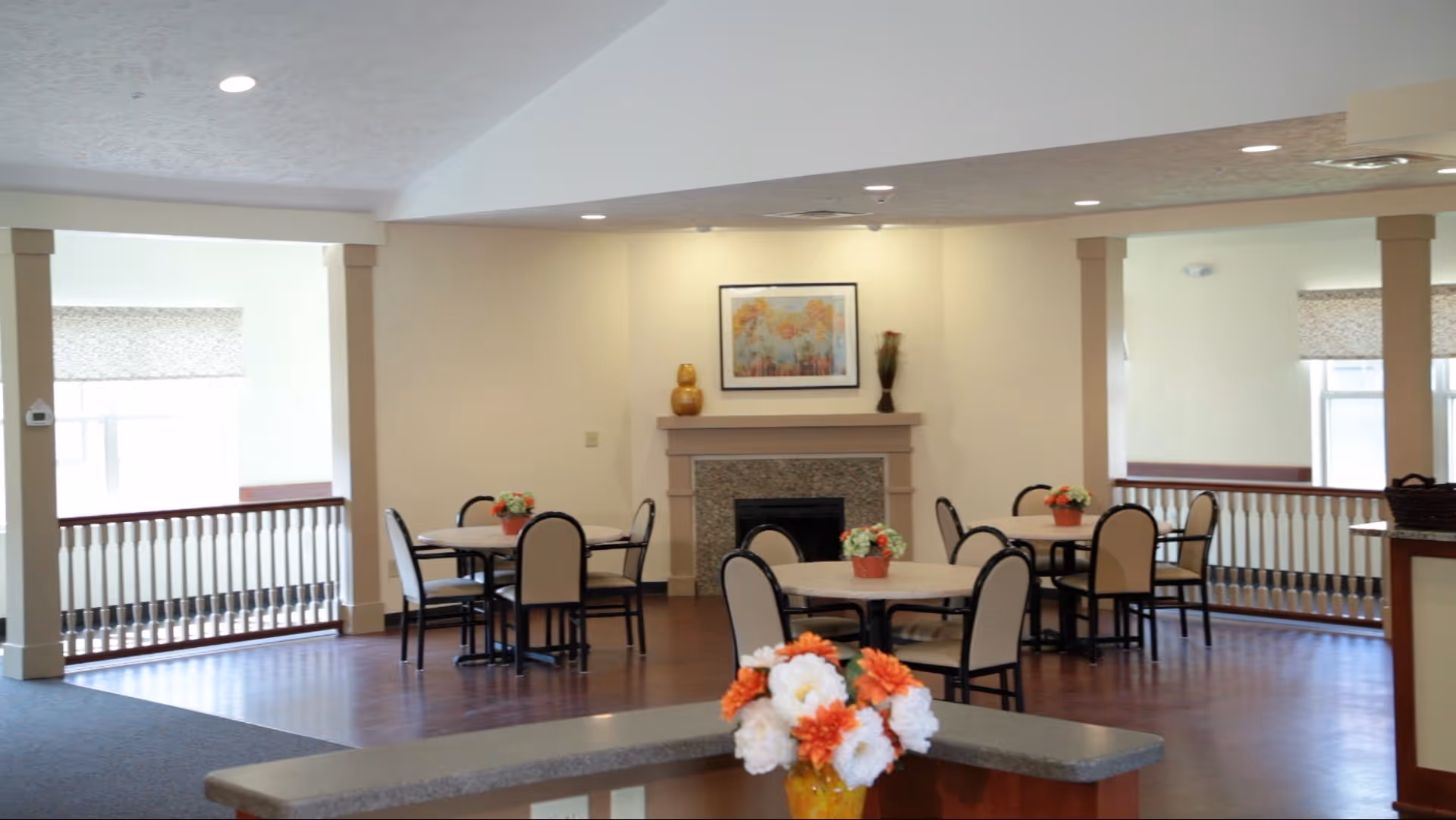 A bright and spacious common area with several round tables and chairs arranged for seating. Each table has a small flower arrangement. The room features a fireplace with decorative items and a framed picture above it. Large windows with patterned blinds allow natural light to fill the space. The area has a combination of carpet and wood flooring and is supported by beige columns.